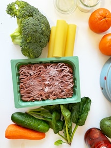 A variety of fresh food items are spread out on a light-colored surface. There is a pack of ground meat in a rectangular container, alongside vegetables like broccoli, spinach, cucumber, and a carrot. Also visible are three cannelloni tubes, two oranges, and jars with some substances inside.
