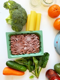 A variety of fresh food items are spread out on a light-colored surface. There is a pack of ground meat in a rectangular container, alongside vegetables like broccoli, spinach, cucumber, and a carrot. Also visible are three cannelloni tubes, two oranges, and jars with some substances inside.