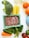 A variety of fresh food items are spread out on a light-colored surface. There is a pack of ground meat in a rectangular container, alongside vegetables like broccoli, spinach, cucumber, and a carrot. Also visible are three cannelloni tubes, two oranges, and jars with some substances inside.