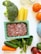 A variety of fresh food items are spread out on a light-colored surface. There is a pack of ground meat in a rectangular container, alongside vegetables like broccoli, spinach, cucumber, and a carrot. Also visible are three cannelloni tubes, two oranges, and jars with some substances inside.