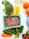 A variety of fresh food items are spread out on a light-colored surface. There is a pack of ground meat in a rectangular container, alongside vegetables like broccoli, spinach, cucumber, and a carrot. Also visible are three cannelloni tubes, two oranges, and jars with some substances inside.