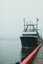 A large fishing boat is docked at a wooden pier on a calm, foggy day. The blue and white vessel features numerous antennas and equipment on its upper deck. The wooden pier in the foreground has a red railing.