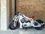 Detailed and futuristic motorcycle with a shiny silver frame and red accents, particularly on the seat and front suspensions. It is positioned next to a large concrete column against a backdrop of a brick wall. The motorcycle has an intricate mechanical design, highlighting various components like the engine and exhaust system.