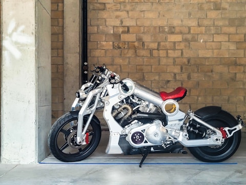 Detailed and futuristic motorcycle with a shiny silver frame and red accents, particularly on the seat and front suspensions. It is positioned next to a large concrete column against a backdrop of a brick wall. The motorcycle has an intricate mechanical design, highlighting various components like the engine and exhaust system.