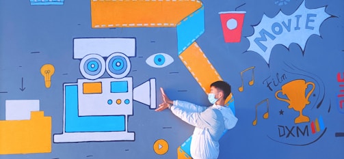 A person wearing a mask and white jacket poses in front of a vibrant mural. The mural includes a large vintage movie camera in blue and orange, an eye, a lightbulb, a cup, and a trophy labeled 'Film DXM'. Musical notes and the word 'MOVIE' are also part of the design.