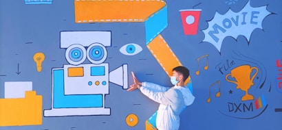 A person wearing a mask and white jacket poses in front of a vibrant mural. The mural includes a large vintage movie camera in blue and orange, an eye, a lightbulb, a cup, and a trophy labeled 'Film DXM'. Musical notes and the word 'MOVIE' are also part of the design.