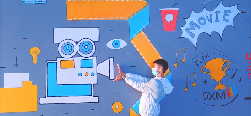 A person wearing a mask and white jacket poses in front of a vibrant mural. The mural includes a large vintage movie camera in blue and orange, an eye, a lightbulb, a cup, and a trophy labeled 'Film DXM'. Musical notes and the word 'MOVIE' are also part of the design.