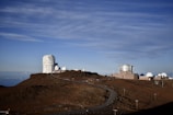 A clear view of an astronomical observatory with multiple large telescopic domes situated on a rocky, elevated terrain. The sky above is overcast with wispy clouds, and the road winding through the site leads up to the domes.