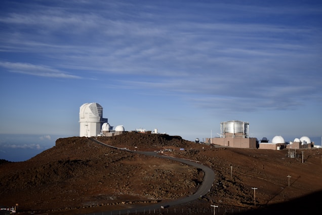 A clear view of an astronomical observatory with multiple large telescopic domes situated on a rocky, elevated terrain. The sky above is overcast with wispy clouds, and the road winding through the site leads up to the domes.