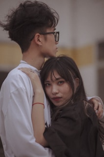 A woman with long, dark hair wearing dark clothing looks into the camera while embracing a man. He has short, curly hair and is wearing glasses and a white shirt. The background is blurred, giving a soft focus to the couple.