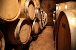 A dimly lit wine cellar filled with large wooden barrels stacked on top of each other. The barrels have metallic rings and markings, and the cellar's earthy walls create a rustic atmosphere.