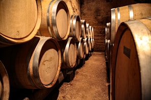 A dimly lit wine cellar filled with large wooden barrels stacked on top of each other. The barrels have metallic rings and markings, and the cellar's earthy walls create a rustic atmosphere.