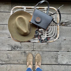 A wicker basket holds an olive green felt hat and a black leather handbag with a decorative scarf. On the weathered wooden floor, a pair of feet in tan shoes and blue jeans are visible.