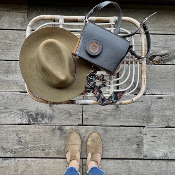 A wicker basket holds an olive green felt hat and a black leather handbag with a decorative scarf. On the weathered wooden floor, a pair of feet in tan shoes and blue jeans are visible.