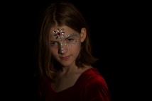 A young person with shoulder-length hair is wearing a red velvet top. Their face is adorned with decorative jewels, including a prominent heart-shaped jewel on the forehead and symmetrical patterns around and above the eyes. The background is dark, highlighting the person's facial expressions and the sparkle of the jewels.