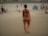 A person wearing a colorful swimsuit walks along a sandy beach. Other beachgoers and scattered beach equipment are visible in the background. The atmosphere appears relaxed with people lounging and socializing.