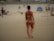 A person wearing a colorful swimsuit walks along a sandy beach. Other beachgoers and scattered beach equipment are visible in the background. The atmosphere appears relaxed with people lounging and socializing.