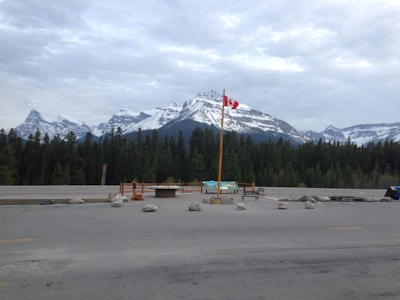 A scenic view featuring a mountain range with snow-capped peaks under a cloudy sky. In the foreground, there is a paved area with a Canadian flag on a tall pole, surrounded by rocks arranged in a circle. The backdrop includes a dense forest of evergreen trees.