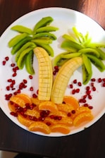 A creative fruit arrangement on a white plate, resembling a tropical scene. Slices of bananas form palm tree trunks, while kiwi slices act as the leaves. Orange segments create a sandy beach effect, surrounded by scattered pomegranate seeds that add a vibrant contrast.