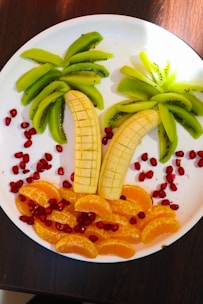 A creative fruit arrangement on a white plate, resembling a tropical scene. Slices of bananas form palm tree trunks, while kiwi slices act as the leaves. Orange segments create a sandy beach effect, surrounded by scattered pomegranate seeds that add a vibrant contrast.