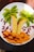 A creative fruit arrangement on a white plate, resembling a tropical scene. Slices of bananas form palm tree trunks, while kiwi slices act as the leaves. Orange segments create a sandy beach effect, surrounded by scattered pomegranate seeds that add a vibrant contrast.