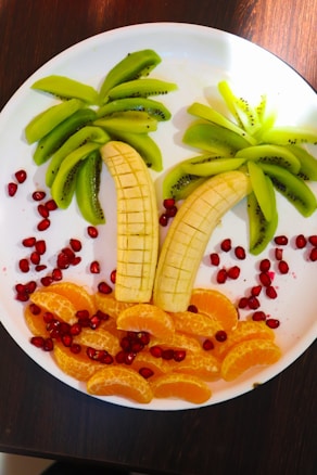 A creative fruit arrangement on a white plate, resembling a tropical scene. Slices of bananas form palm tree trunks, while kiwi slices act as the leaves. Orange segments create a sandy beach effect, surrounded by scattered pomegranate seeds that add a vibrant contrast.