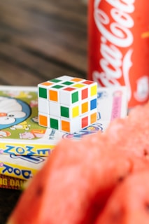 A partially solved Rubik's cube is placed on a colorful comic book. In the background, there is a red Coca-Cola can partially visible. The image also features slices of watermelon in the foreground.
