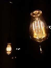 Two illuminated Edison-style light bulbs create a warm and ambient glow against a dark background. The bulbs highlight their intricate filaments, casting subtle reflections.