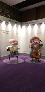 Two cartoonish pig figures are displayed standing on a carpeted purple floor. One is playing a French horn and wearing glasses, while the other is playing a cello. The backdrop features text reading 'Hong Kong Sinfonietta' and 'see the music.' The background is mainly white and gray.