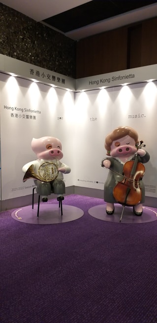 Two cartoonish pig figures are displayed standing on a carpeted purple floor. One is playing a French horn and wearing glasses, while the other is playing a cello. The backdrop features text reading 'Hong Kong Sinfonietta' and 'see the music.' The background is mainly white and gray.