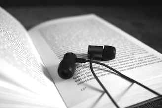 A pair of black in-ear headphones resting on an open book. The book's text is in focus and readable, while the background is blurred, drawing attention to the headphones and the pages.