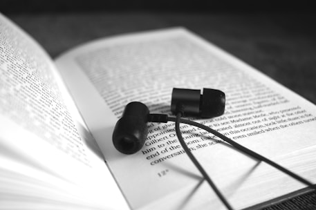 A pair of black in-ear headphones resting on an open book. The book's text is in focus and readable, while the background is blurred, drawing attention to the headphones and the pages.