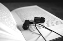 A pair of black in-ear headphones resting on an open book. The book's text is in focus and readable, while the background is blurred, drawing attention to the headphones and the pages.