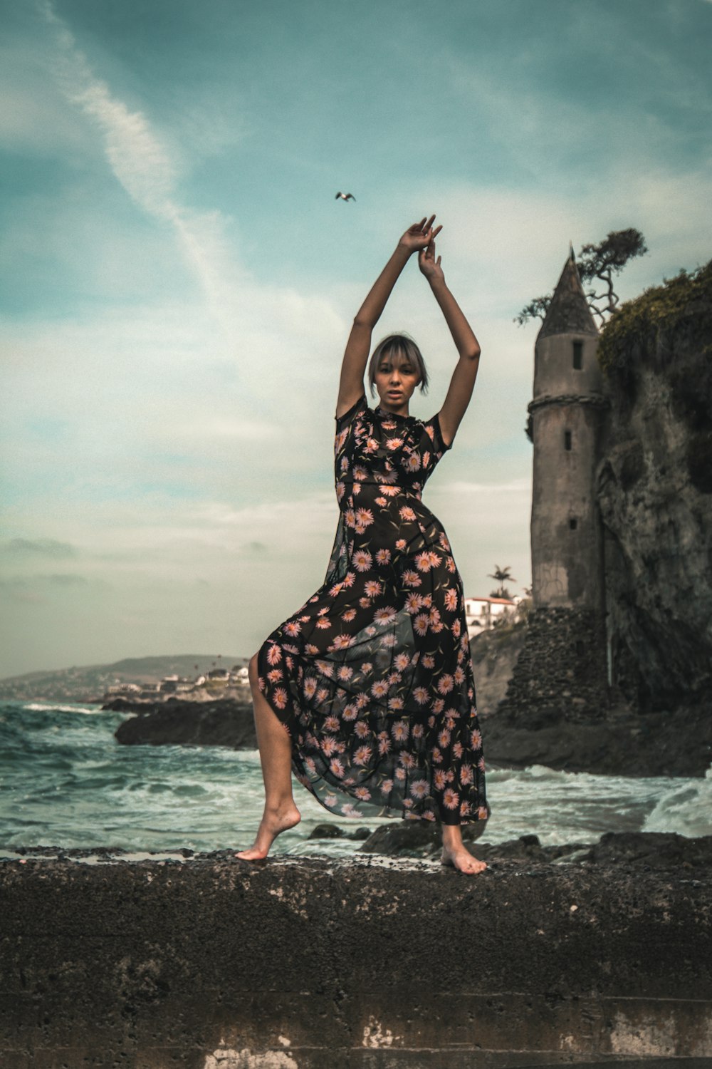 woman in black and white floral dress standing on gray rock formation near body of water