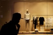 A storefront display featuring an arrangement of clothing items on mannequin and racks. A mannequin is dressed in a light-colored hoodie and sweatpants, surrounded by folded and hanging clothes. The background consists of a plain-textured wall with signage promoting sustainably sourced cotton. The silhouette of a person in a hat is visible in the foreground.