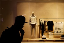A storefront display featuring an arrangement of clothing items on mannequin and racks. A mannequin is dressed in a light-colored hoodie and sweatpants, surrounded by folded and hanging clothes. The background consists of a plain-textured wall with signage promoting sustainably sourced cotton. The silhouette of a person in a hat is visible in the foreground.