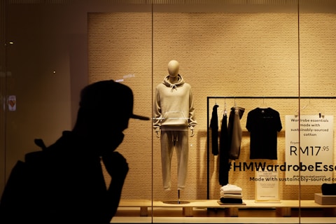 A storefront display featuring an arrangement of clothing items on mannequin and racks. A mannequin is dressed in a light-colored hoodie and sweatpants, surrounded by folded and hanging clothes. The background consists of a plain-textured wall with signage promoting sustainably sourced cotton. The silhouette of a person in a hat is visible in the foreground.