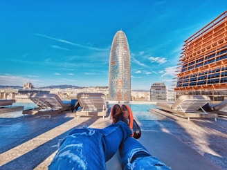A perspective from a rooftop pool area overlooking a cityscape with a distinctive modern skyscraper in the center. The sky is bright and clear, providing a vivid contrast to the buildings. Sun loungers are placed along the poolside, and a person’s legs are visible, suggesting a relaxed atmosphere.