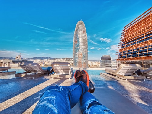 A perspective from a rooftop pool area overlooking a cityscape with a distinctive modern skyscraper in the center. The sky is bright and clear, providing a vivid contrast to the buildings. Sun loungers are placed along the poolside, and a person’s legs are visible, suggesting a relaxed atmosphere.