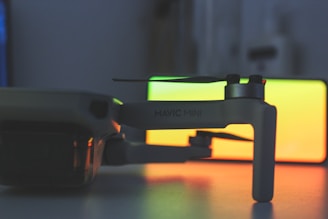 A close-up view of a small drone, specifically a DJI Mavic Mini, with a blurred, colorful background displaying a gradient of green and orange hues. The focus is on the drone's arm and propeller, with the brand name 'Mavic Mini' visible on the side.