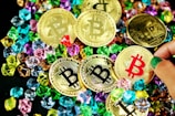 A collection of gold coins featuring the Bitcoin logo is scattered among colorful gemstones. The coins have black, red, and gold bitcoin symbols, while various hands can be seen interacting with them.