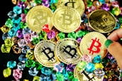 A collection of gold coins featuring the Bitcoin logo is scattered among colorful gemstones. The coins have black, red, and gold bitcoin symbols, while various hands can be seen interacting with them.