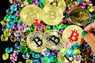 A collection of gold coins featuring the Bitcoin logo is scattered among colorful gemstones. The coins have black, red, and gold bitcoin symbols, while various hands can be seen interacting with them.