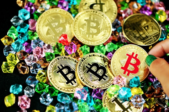 A collection of gold coins featuring the Bitcoin logo is scattered among colorful gemstones. The coins have black, red, and gold bitcoin symbols, while various hands can be seen interacting with them.