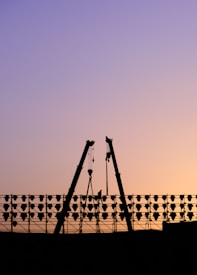 Silhouette of a construction site at sunset with two cranes positioned in an A-frame structure. A worker stands on scaffolding amidst a series of lights or hoisting equipment. The sky transitions from purple to a soft orange, indicating either dawn or dusk.