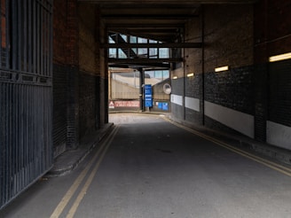 An industrial alleyway with brick walls on both sides and overhead structures casting shadows. The passageway leads to a bright area with road signs, including a speed limit warning of 4 miles per hour. Yellow double lines run along the edges of the road, and there are pipes and metal fixtures attached to the walls.