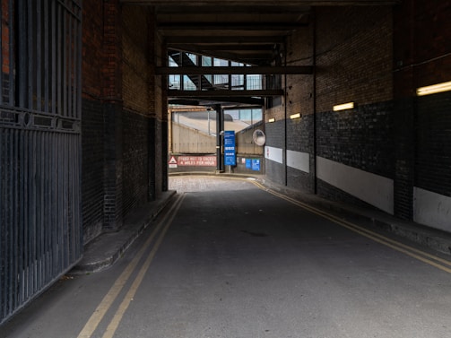 An industrial alleyway with brick walls on both sides and overhead structures casting shadows. The passageway leads to a bright area with road signs, including a speed limit warning of 4 miles per hour. Yellow double lines run along the edges of the road, and there are pipes and metal fixtures attached to the walls.