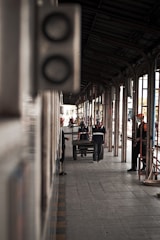 A perspective view of a railway station platform, showing people at work. The platform is covered with a metal roof supported by arches, providing a sense of depth. Several workers wearing uniforms and safety gear are engaged in various activities. A person is leaning against the wall on the left, and another is pushing a trolley further down the platform. The scene conveys a sense of routine and activity.