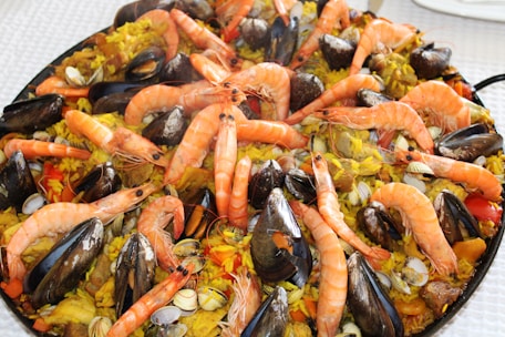 A large dish of seafood paella, featuring beautifully arranged shrimp, mussels, clams, and pieces of chicken on a bed of saffron-infused rice with visible pieces of red and yellow peppers. The ingredients are vibrant and placed in a circular pattern, creating an appetizing presentation.
