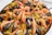 A large dish of seafood paella, featuring beautifully arranged shrimp, mussels, clams, and pieces of chicken on a bed of saffron-infused rice with visible pieces of red and yellow peppers. The ingredients are vibrant and placed in a circular pattern, creating an appetizing presentation.